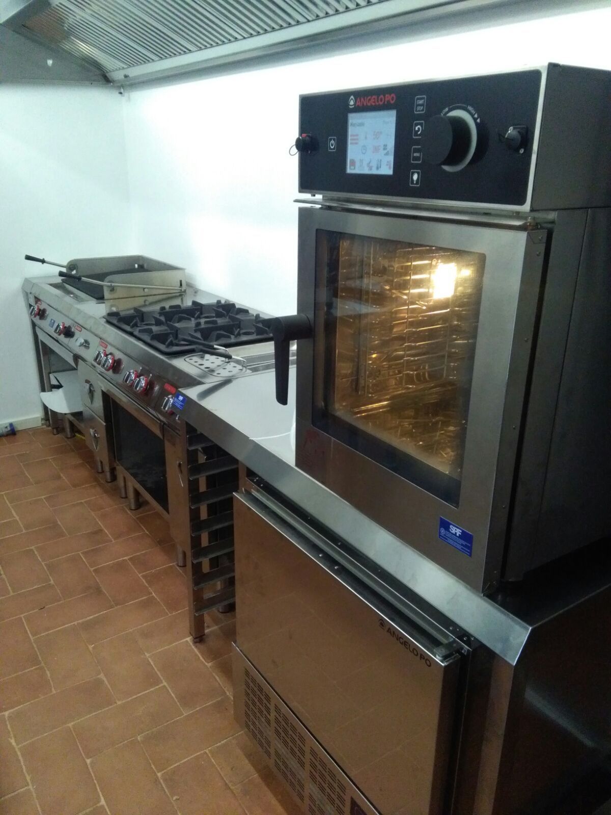 Cucina professionale con piano cottura, forno e tavolo di lavoro in acciaio inox lungo una parete piastrellata.