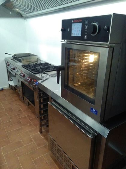 Cucina professionale con forno, piano cottura e bancone in acciaio inox lungo una parete piastrellata.