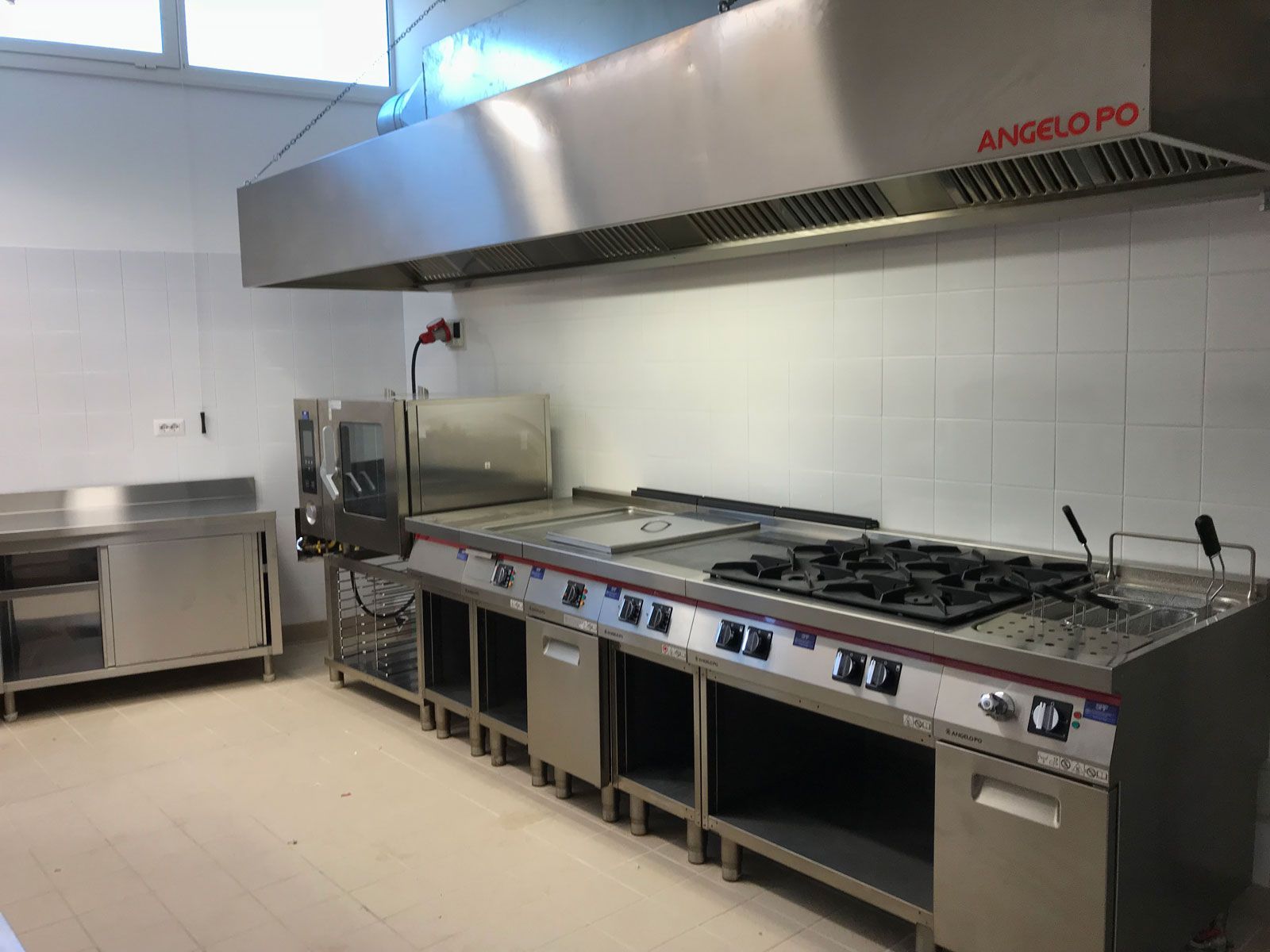 Cucina professionale con piano cottura in acciaio inox, cappa aspirante e banconi di lavoro addossati a pareti piastrellate.