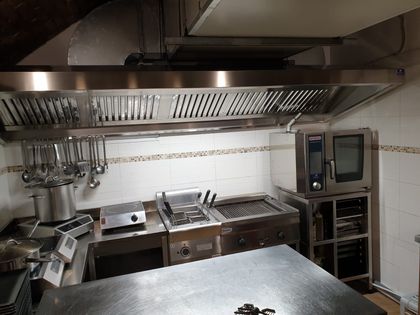 Cucina professionale con bancone di lavoro in acciaio inox, pentole sospese, forno e ampia cappa aspirante.