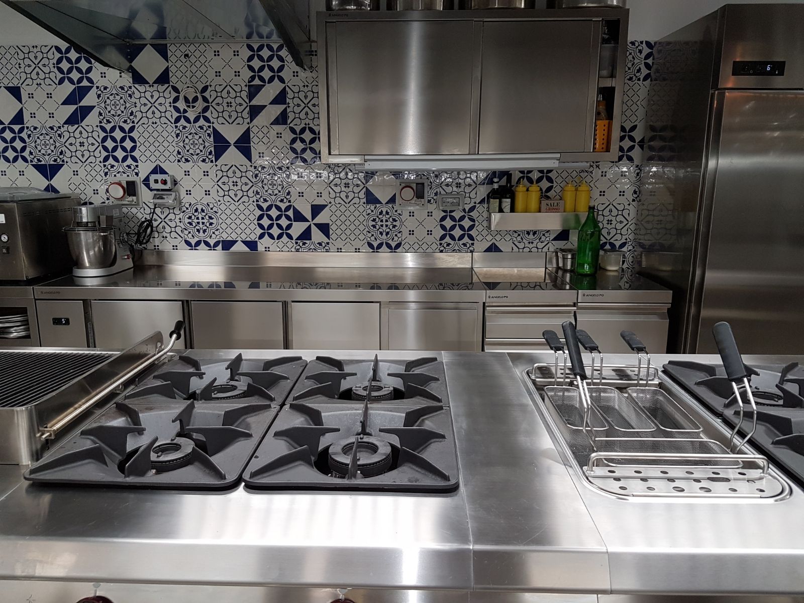 Cucina in acciaio inox con piano cottura a gas, utensili e paraschizzi piastrellato in blu e bianco.
