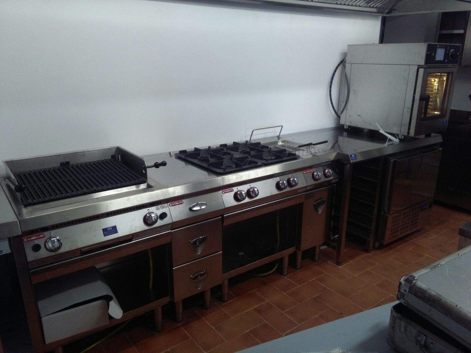Cucina professionale con piano cottura a gas in acciaio inox, griglia e bancone di lavoro, pavimento e pareti piastrellati.
