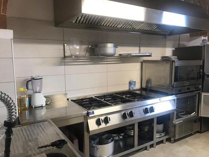 Cucina professionale con piano cottura, cappa e attrezzature per la preparazione dei cibi in acciaio inox lungo una parete piastrellata.
