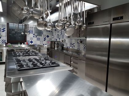 Cucina professionale moderna in acciaio inox con pentole sospese, piano cottura a gas e grande frigorifero.