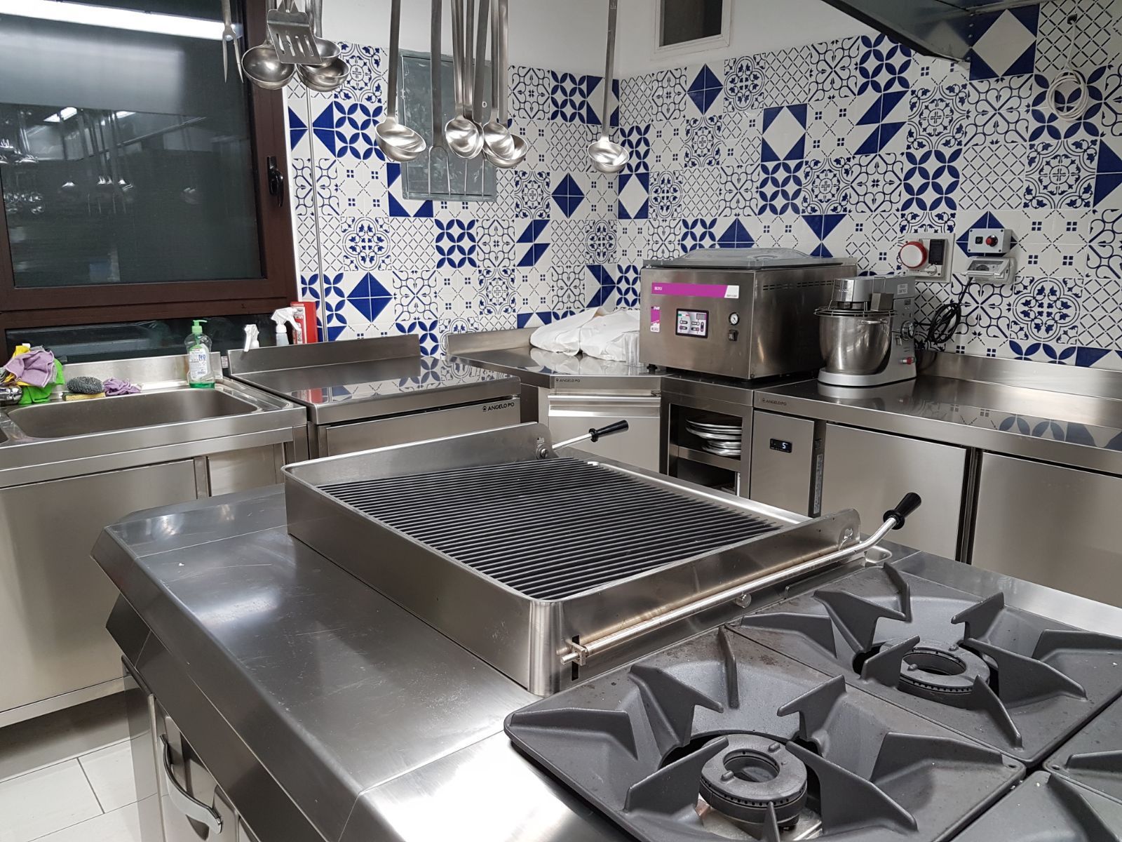 Cucina professionale con ripiani in acciaio inox, piano cottura a gas e rivestimento a piastrelle con motivo blu e bianco.