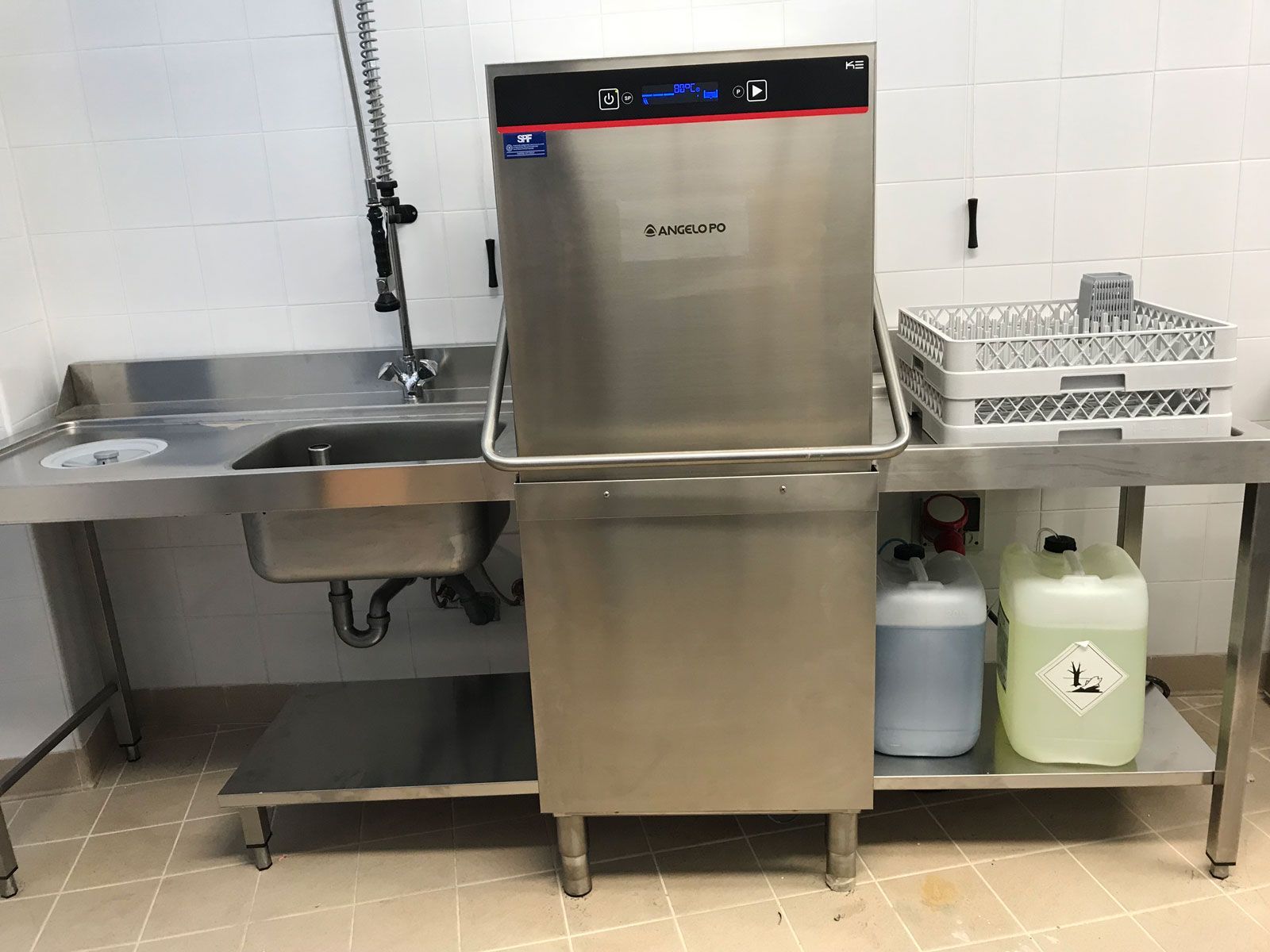 Lavello e lavastoviglie professionali in acciaio inox in un locale di servizio piastrellato, con i prodotti per la pulizia riposti al di sotto.