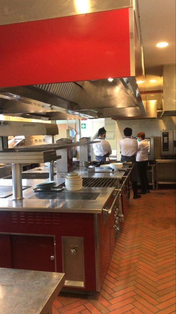 Cucina professionale con banconi in acciaio inox, due addetti ai fornelli e pavimento in piastrelle rosse.