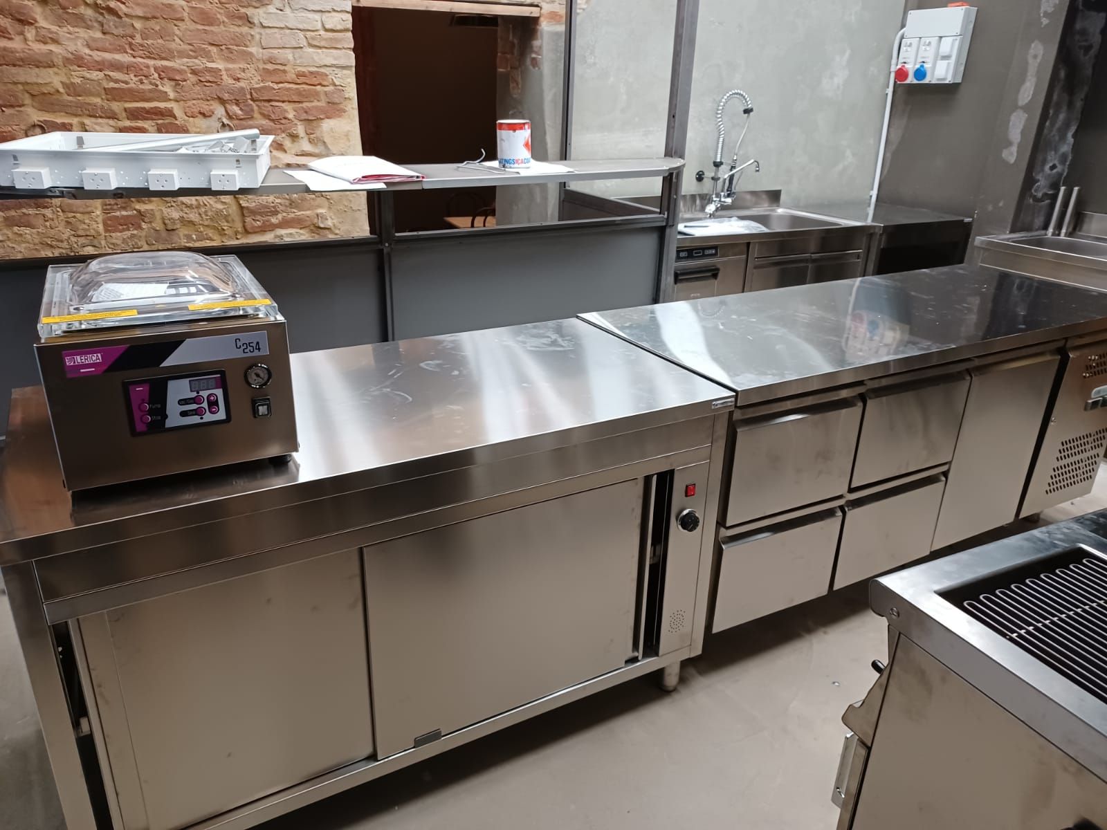 Bancone da cucina professionale in acciaio inox con ripiani, lavello e contenitori nella zona di preparazione.