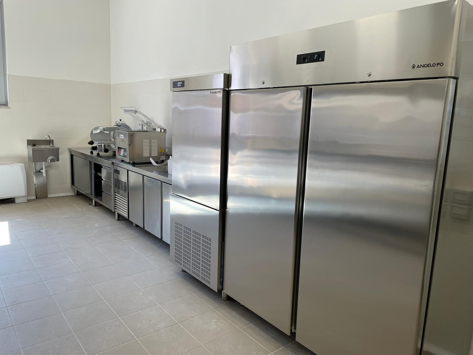 Cucina professionale con ripiani in acciaio inox e grandi frigoriferi lungo una parete piastrellata.
