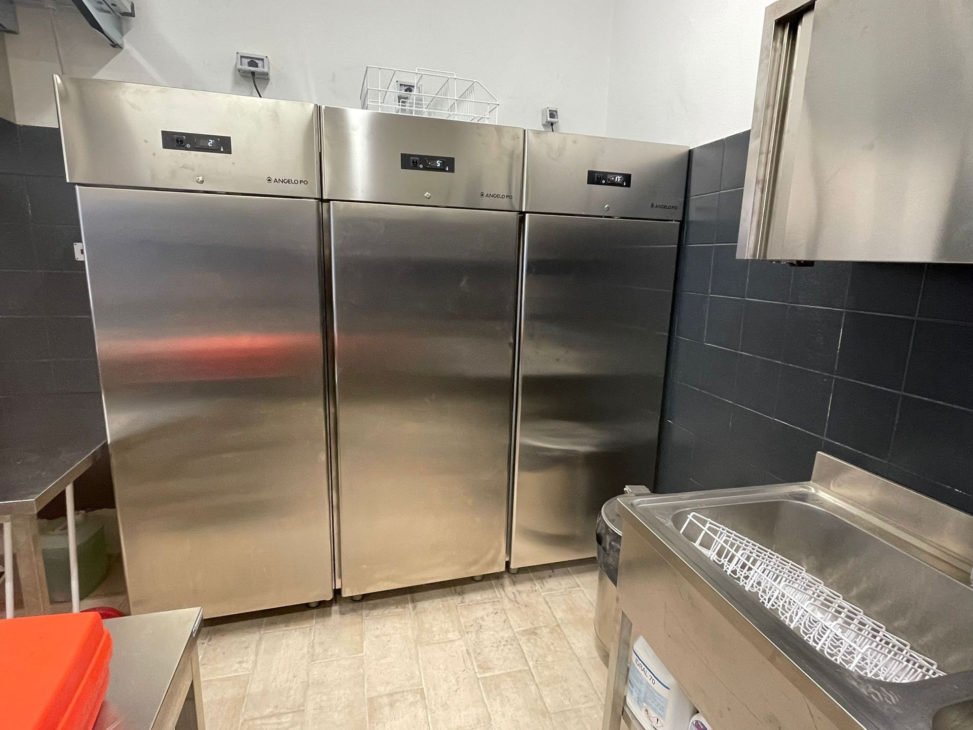 Cucina professionale con tre frigoriferi in acciaio inox, pareti rivestite con piastrelle scure e bancone con lavello in metallo.