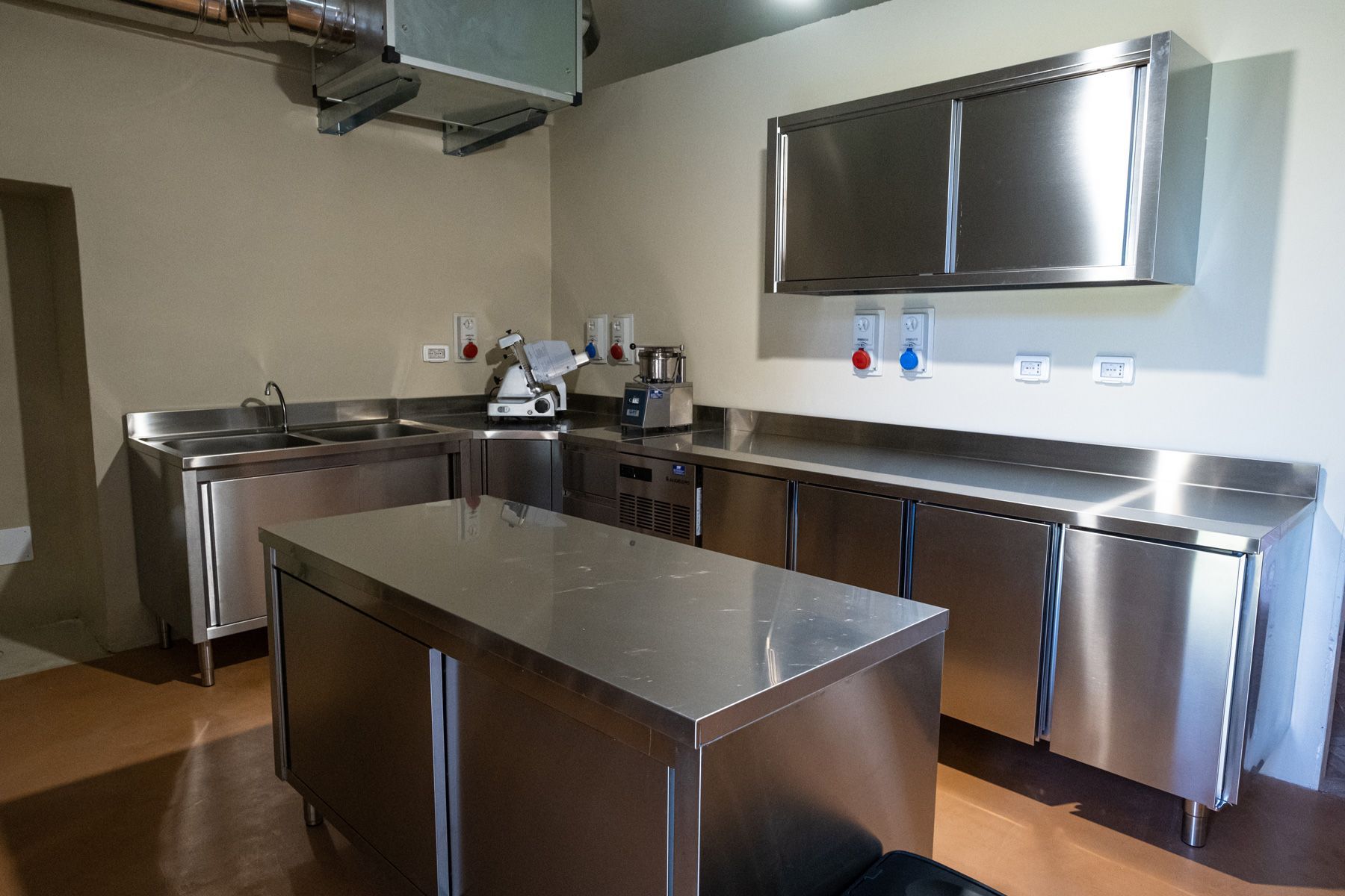 Cucina professionale in acciaio inox con isola di preparazione, banconi, lavelli e pensili a muro.