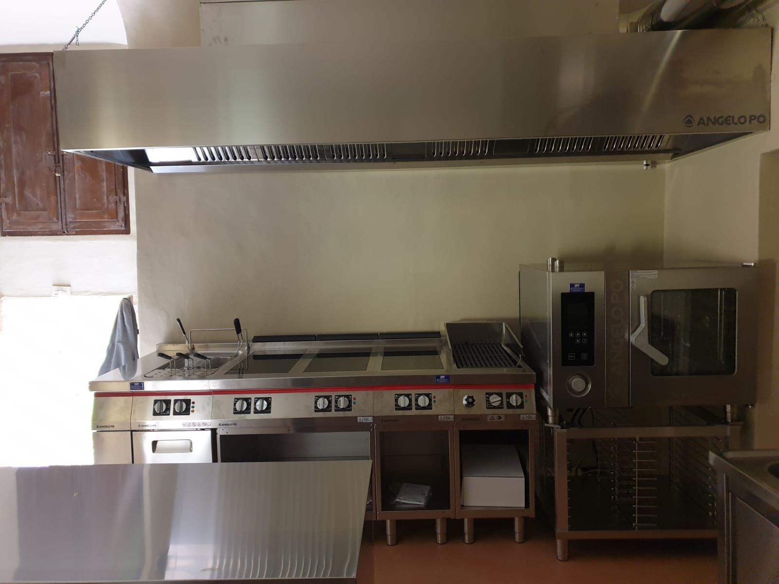 Cucina professionale con piano cottura, forni e grande cappa aspirante in acciaio inox lungo una parete beige.