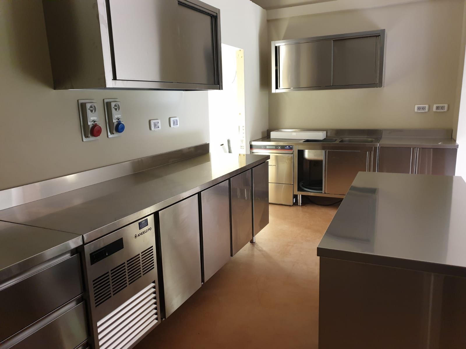 Cucina commerciale vuota in acciaio inox con banconi, armadietti, lavello e contenitori a parete.