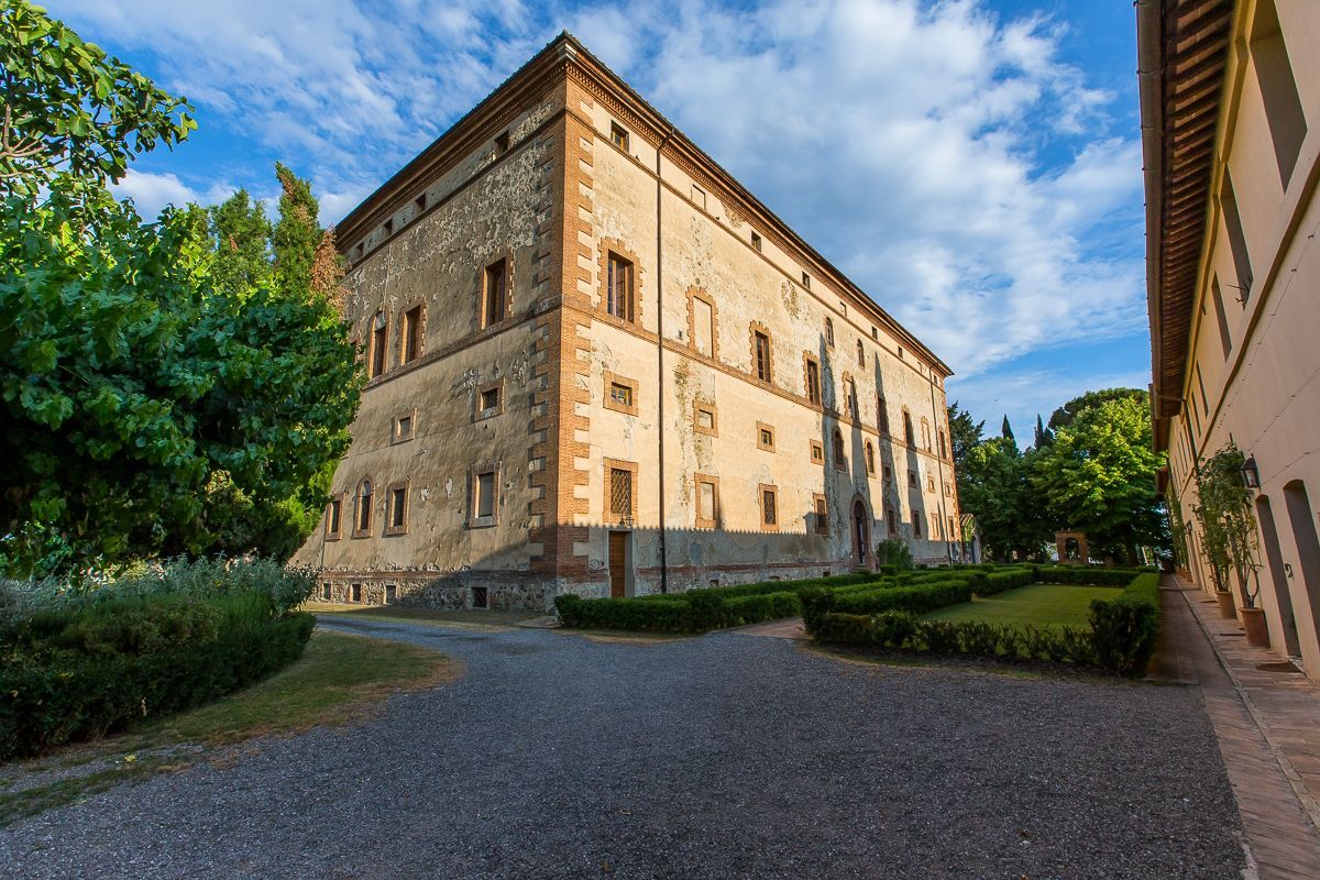 Edificio storico in pietra con finestre ad arco e cortile interno, sotto un cielo azzurro con alberi nelle vicinanze.