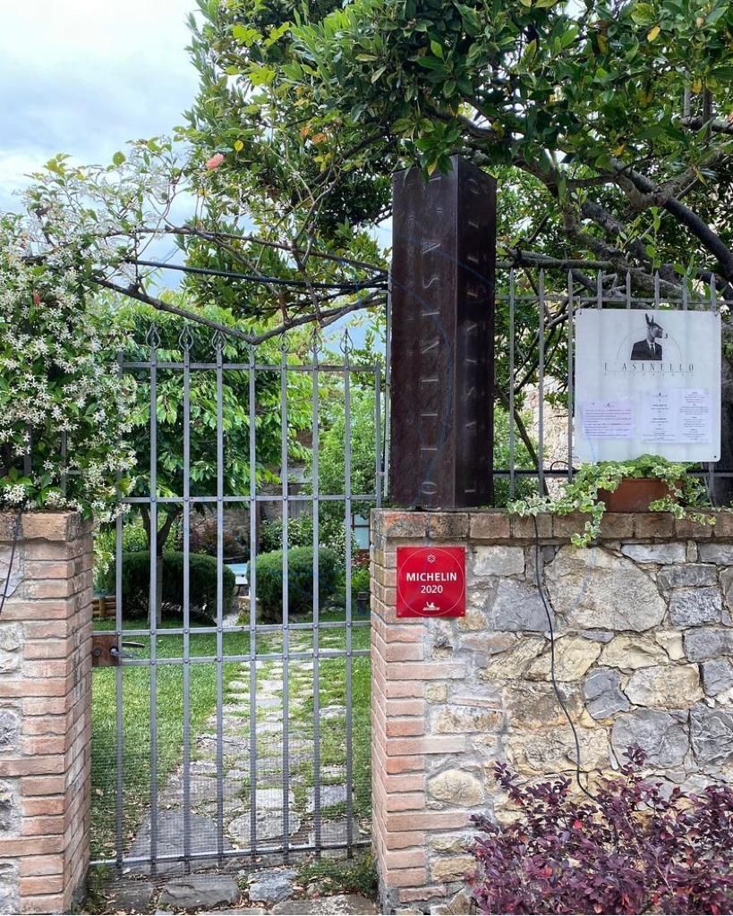 Ingresso con cancello in metallo e muro in pietra con cartello, circondato da vegetazione e alberi.