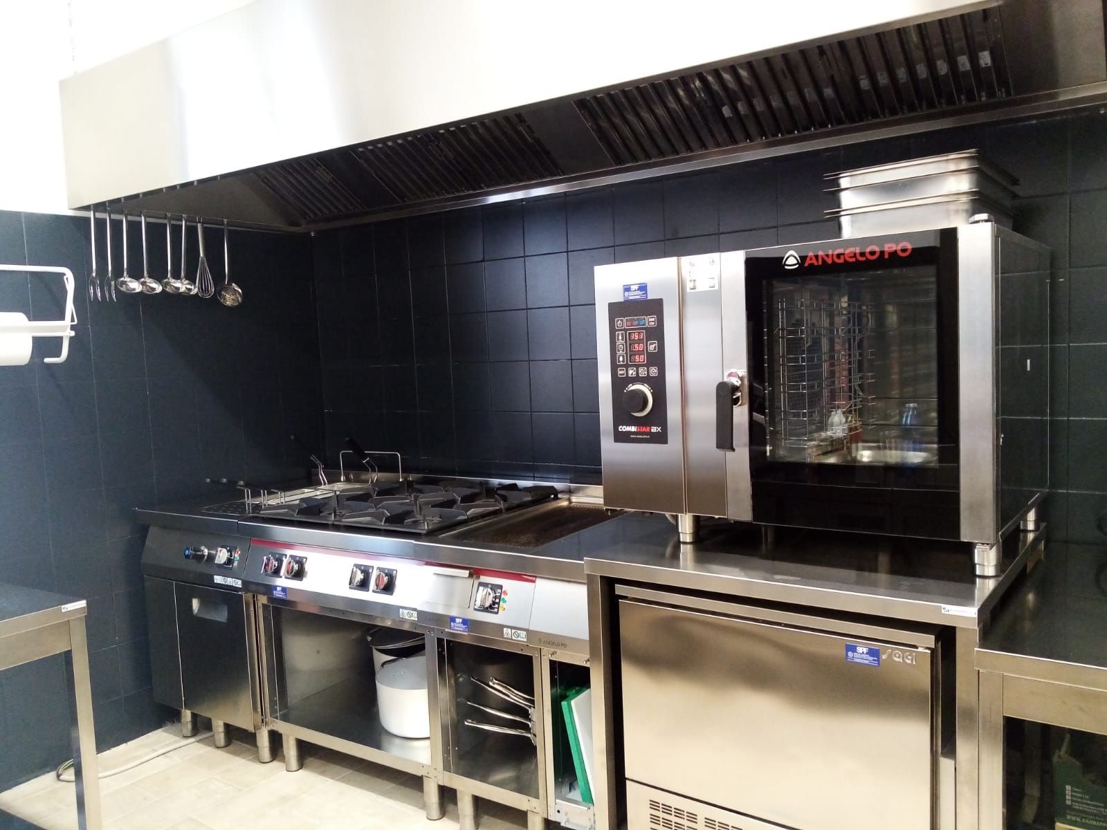 Cucina professionale con piano cottura, forno e grandi attrezzature industriali in acciaio inox, addossata a una parete scura.