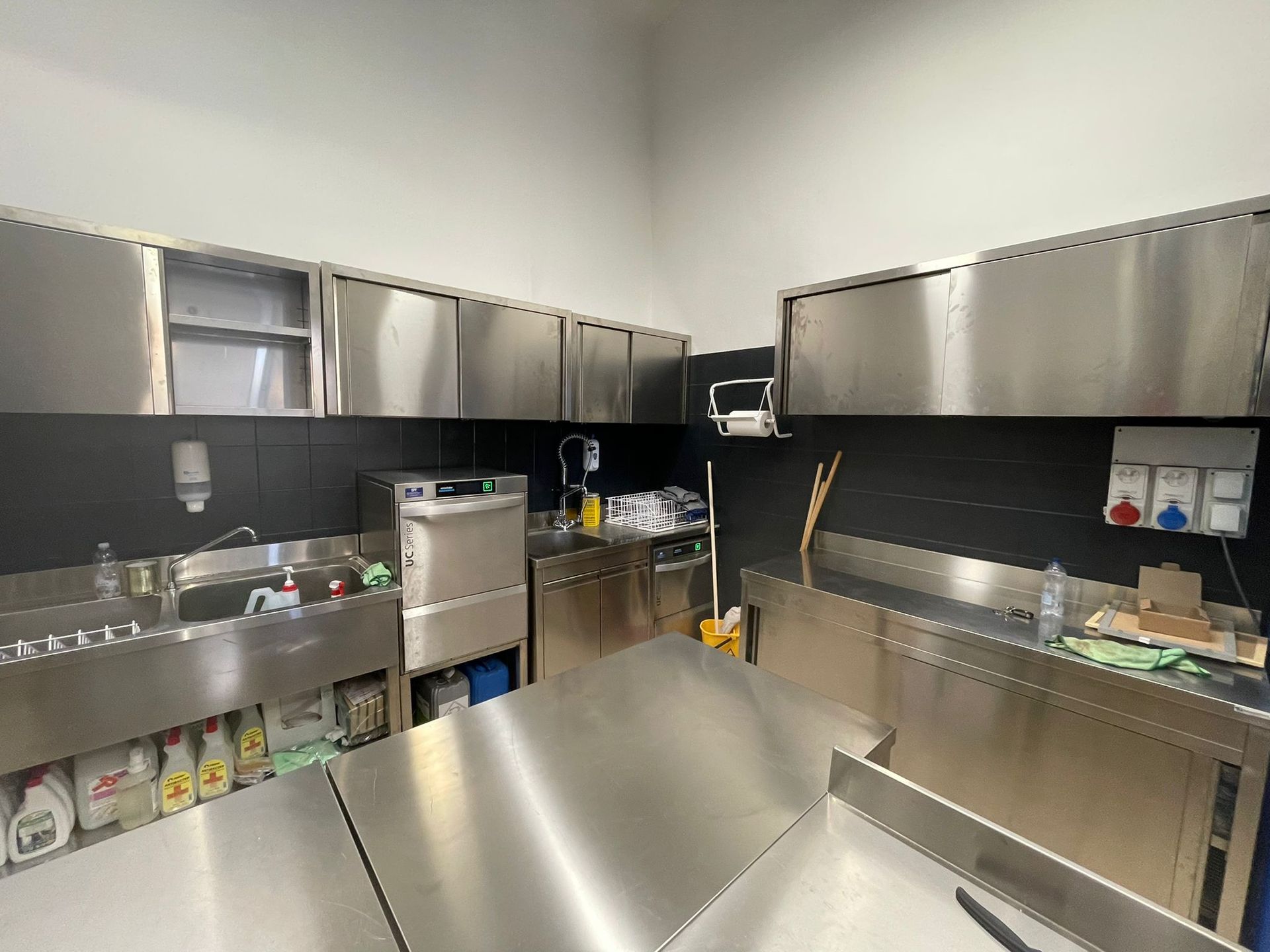 Cucina professionale in acciaio inox con lavelli, banconi e pensili in una zona di preparazione pulita.