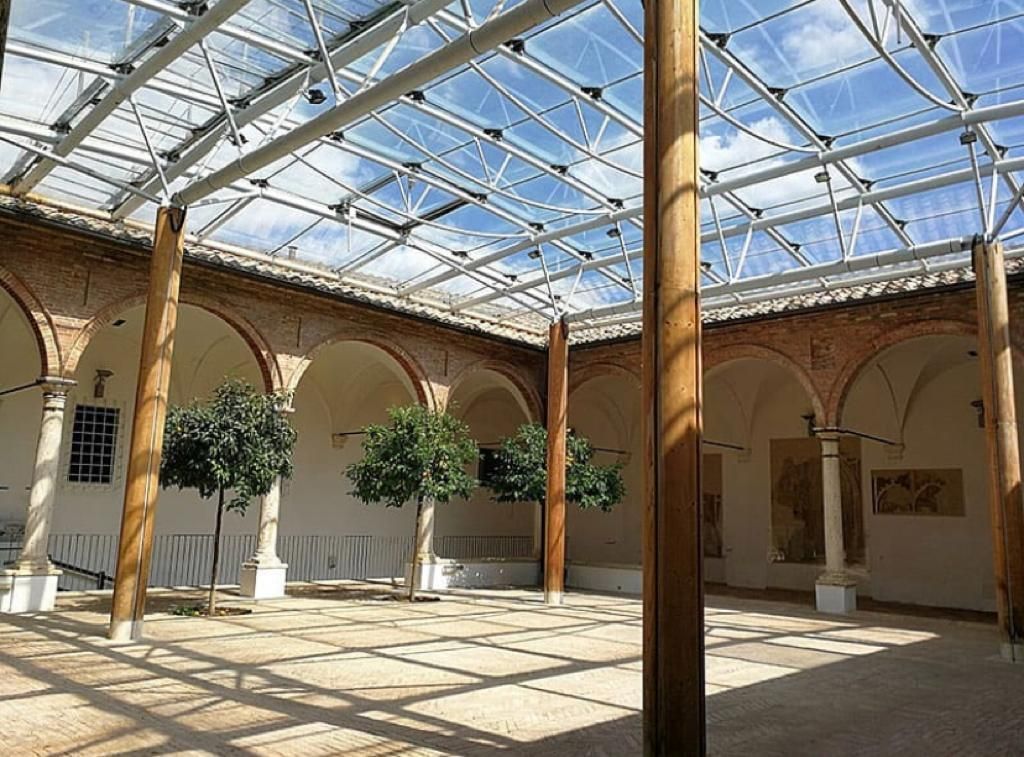 Cortile a cielo aperto con tetto in vetro, pilastri in legno, archi e alberi in vaso baciati dal sole.