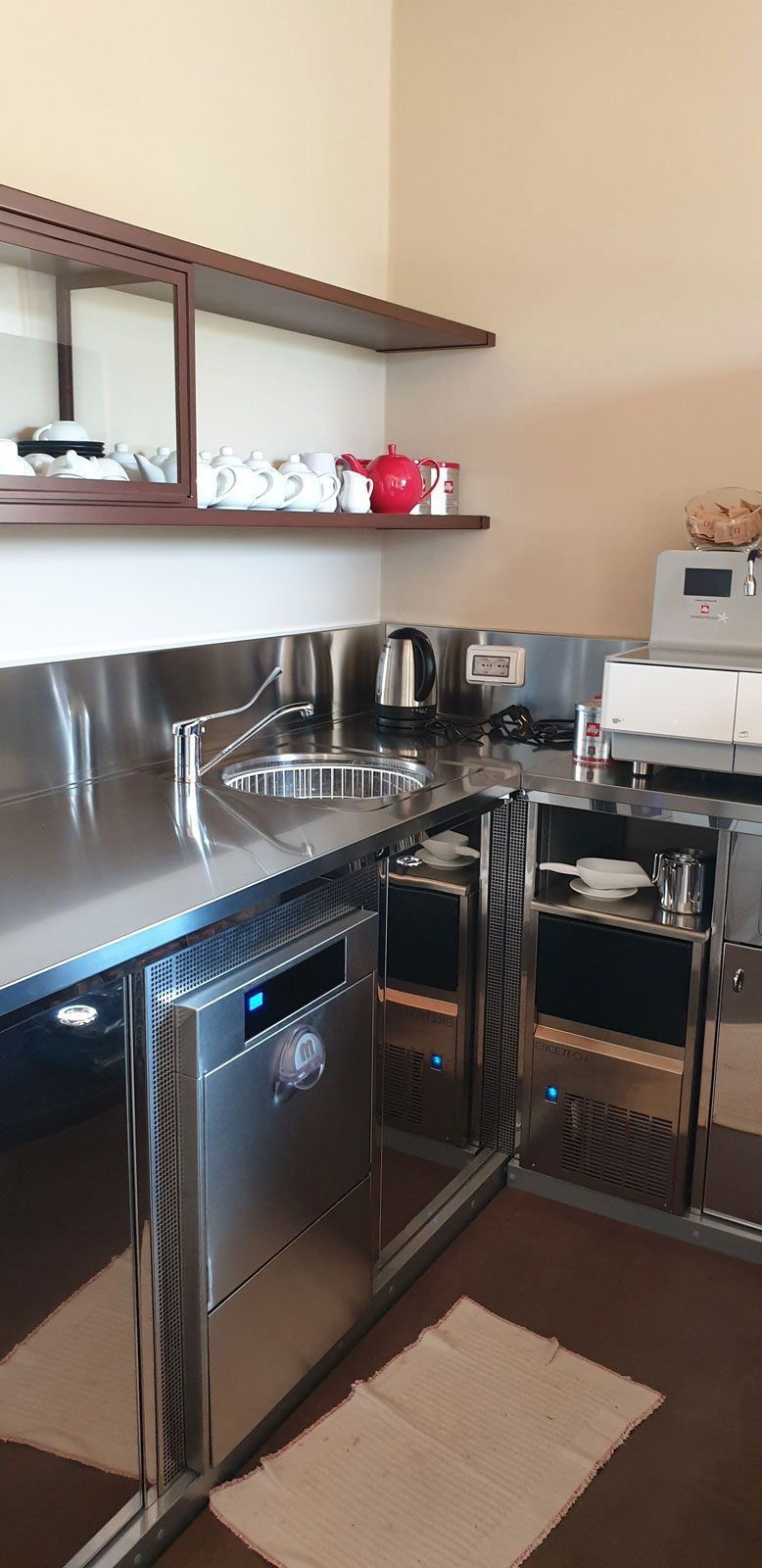 Angolo cucina compatto con lavello in acciaio inox, forno a microonde, bollitore, lavastoviglie e tazze rosse su uno scaffale.