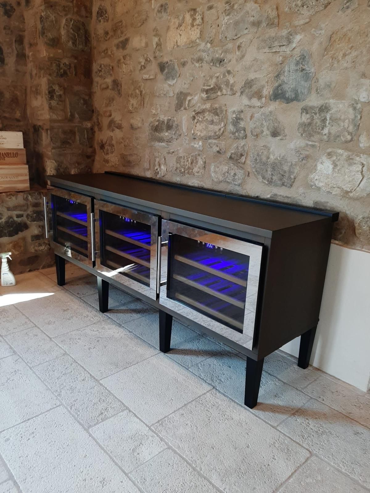Mobile porta TV lungo e nero con ante in vetro e luci blu, addossato a una parete in pietra su un pavimento piastrellato.
