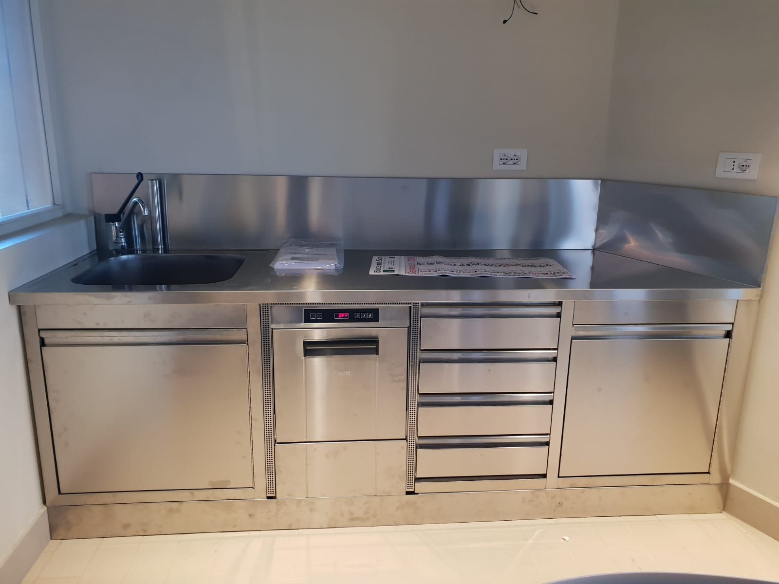 Bancone da cucina integrato in acciaio inox con lavello, piano cottura, forno e cassetti, sotto un rivestimento piastrellato.