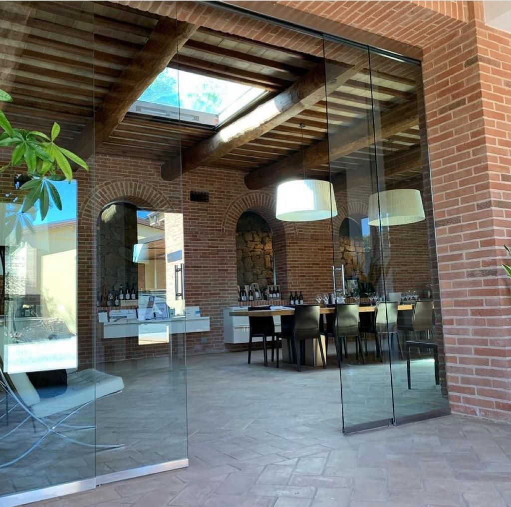 Zona pranzo esterna in mattoni con porte a vetri, travi in legno a vista e un lungo tavolo all'interno.