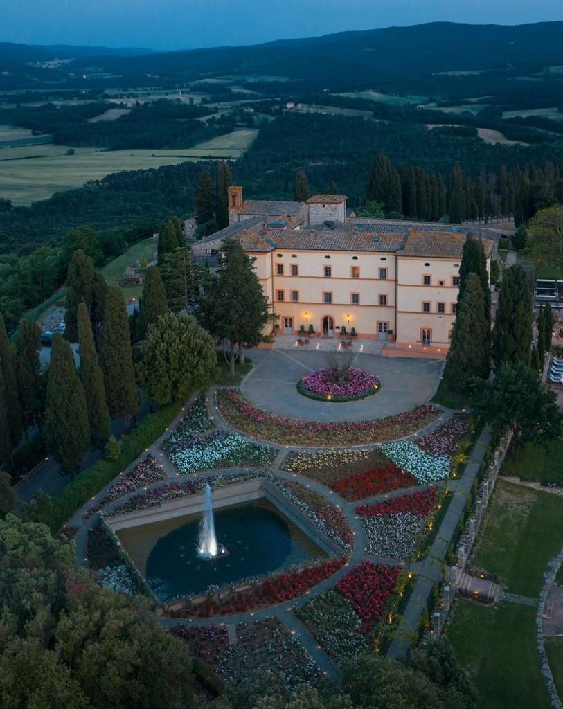 Veduta aerea di una villa illuminata con giardini, fontana e piscina al crepuscolo, circondata da campagna e colline.