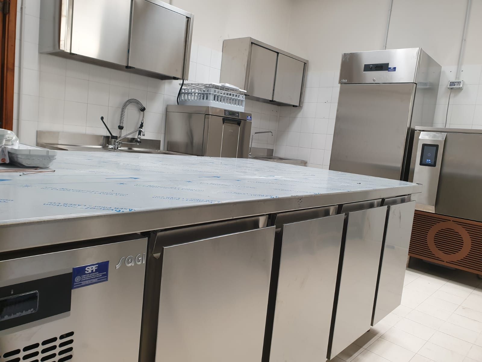 Cucina professionale con ripiani in acciaio inox, lavello, armadietti e un grande frigorifero, il tutto illuminato da una luce intensa.