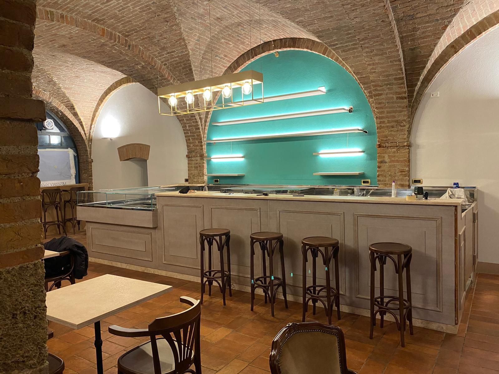 Bar-caffetteria con archi in mattoni, scaffali retroilluminati color turchese, bancone bianco e sgabelli in legno.