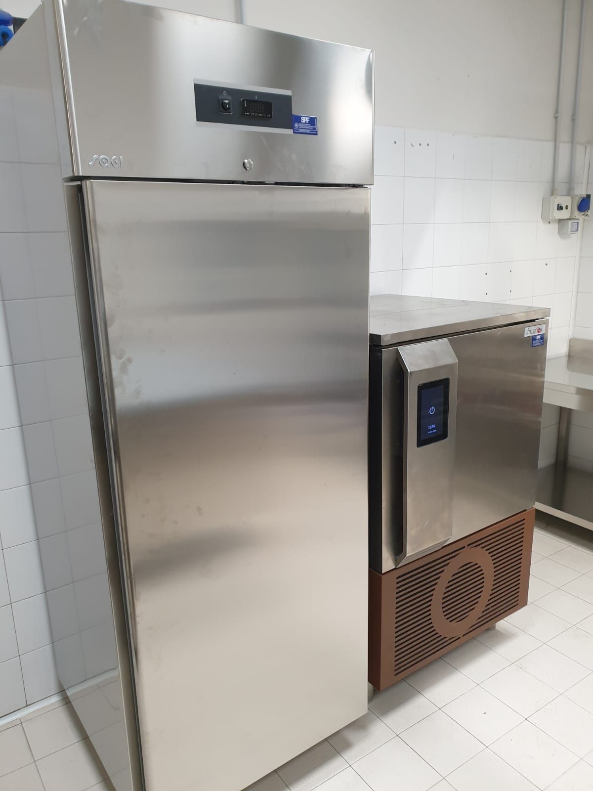 Frigorifero commerciale in acciaio inox e macchina per il ghiaccio in una cucina piastrellata di bianco.