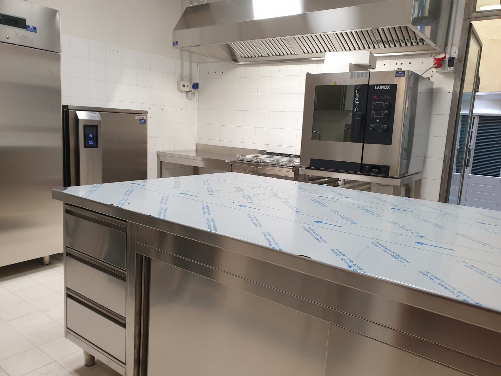 Cucina professionale in acciaio inox con tavoli da lavoro, frigorifero, forno e un'ampia isola rivestita con pellicola blu.
