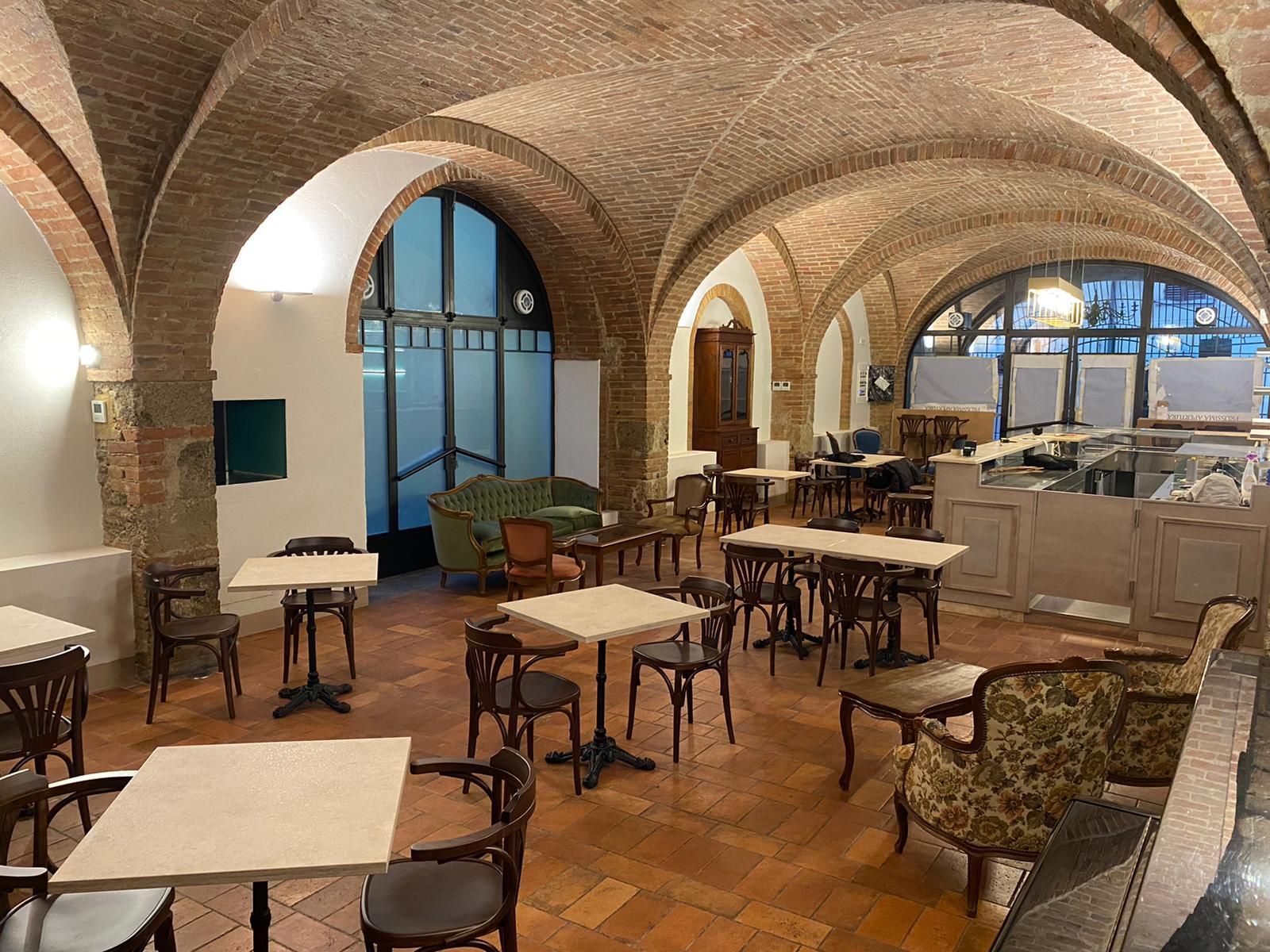 Interno di un caffè con facciata ad arco in mattoni, tavoli e sedie vuoti e una calda illuminazione soffusa.