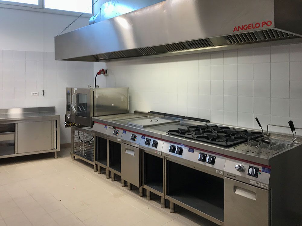 Cucina professionale in acciaio inox con piano cottura a gas, cappa aspirante e tavoli da lavoro in un locale piastrellato.