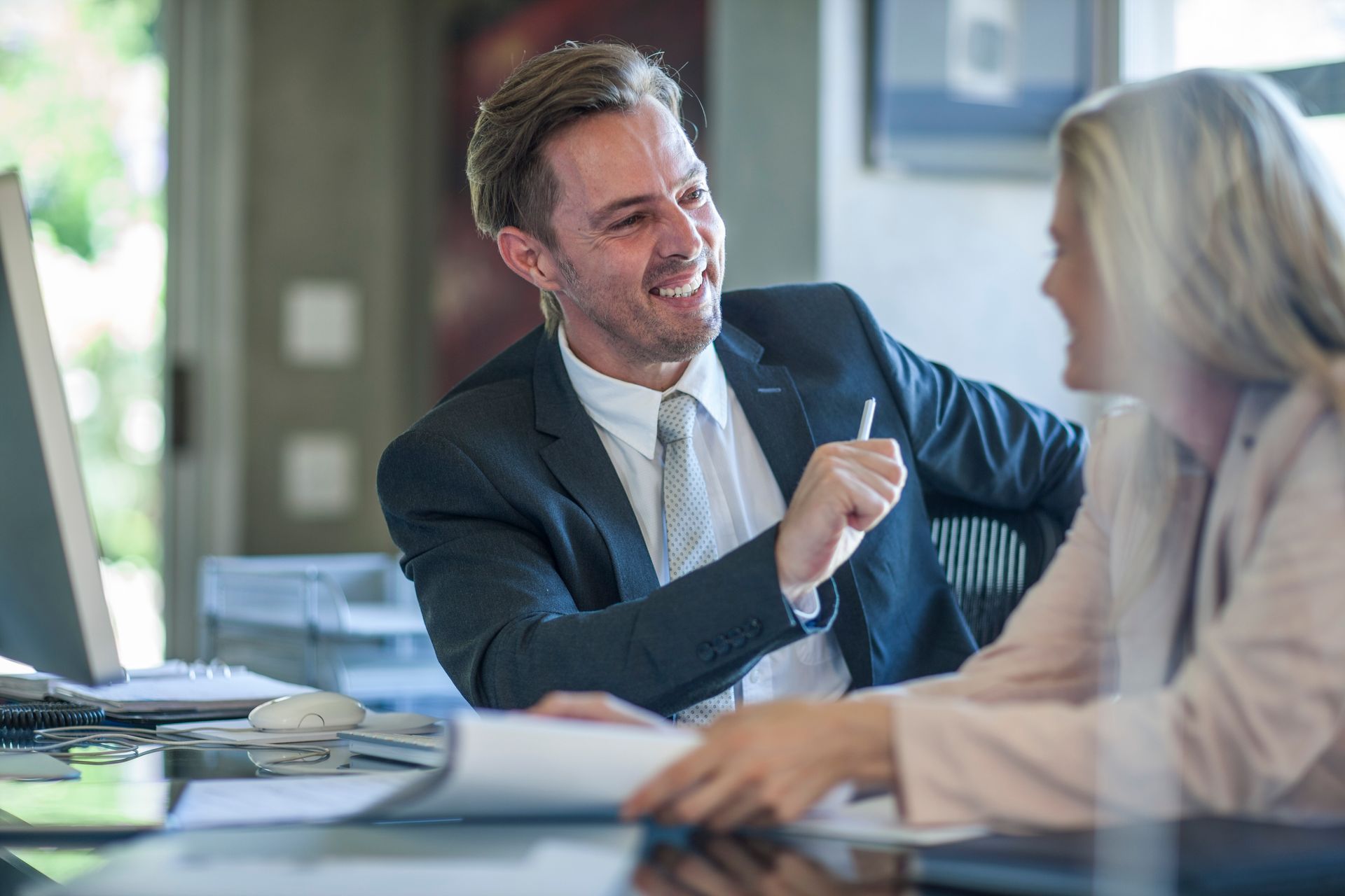 Man in suit laughs, gesturing with pen at woman holding papers, in office.