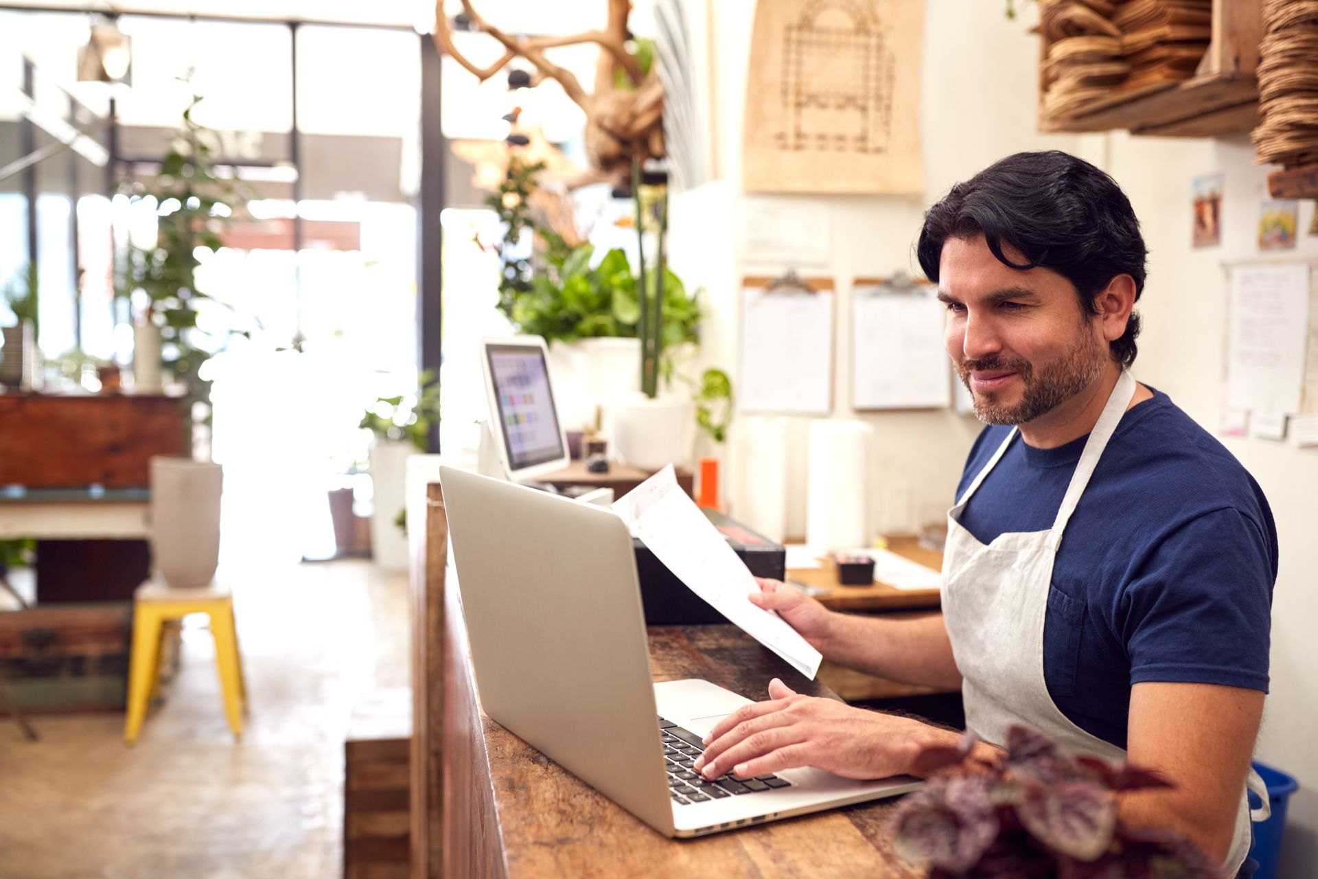 Man in apron uses laptop at a shop counter, holding papers, smiling.