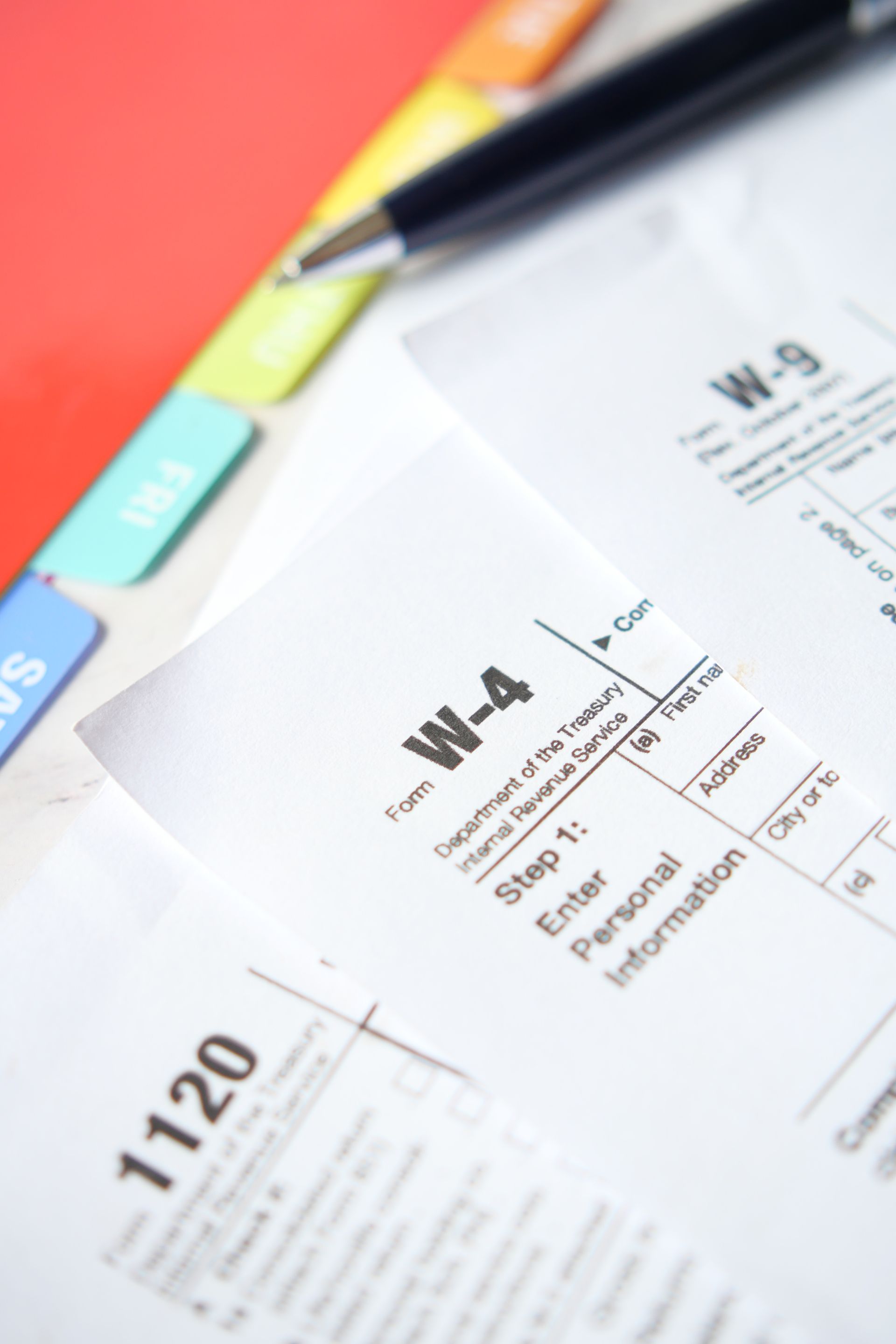 W-4 and W-9 tax forms with a pen, file tabs, and a red folder in the background.