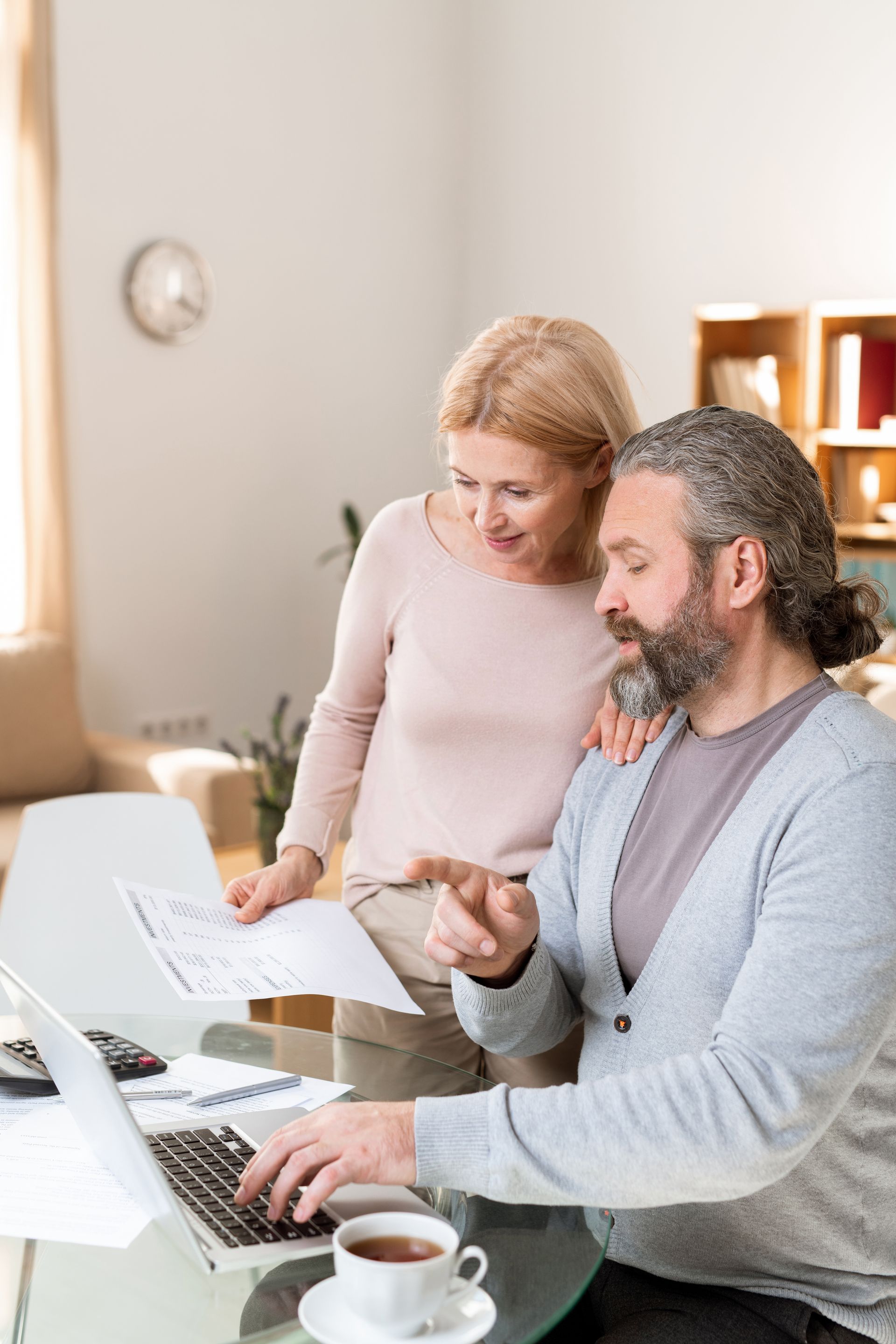 Couple reviewing documents and using a laptop, one points, in a bright home setting.