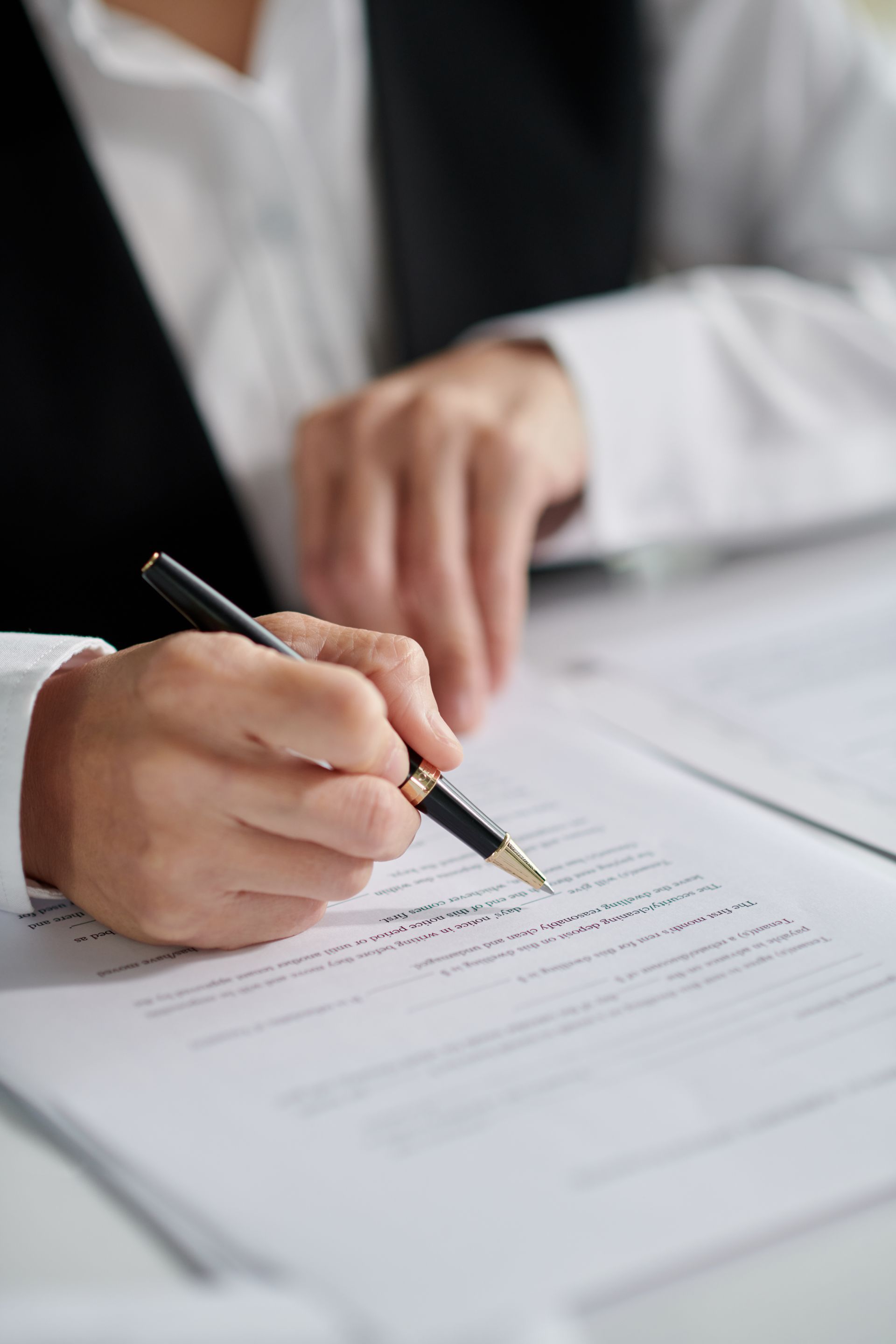 Person signing a document with a pen at a desk, wearing a white shirt and black vest.