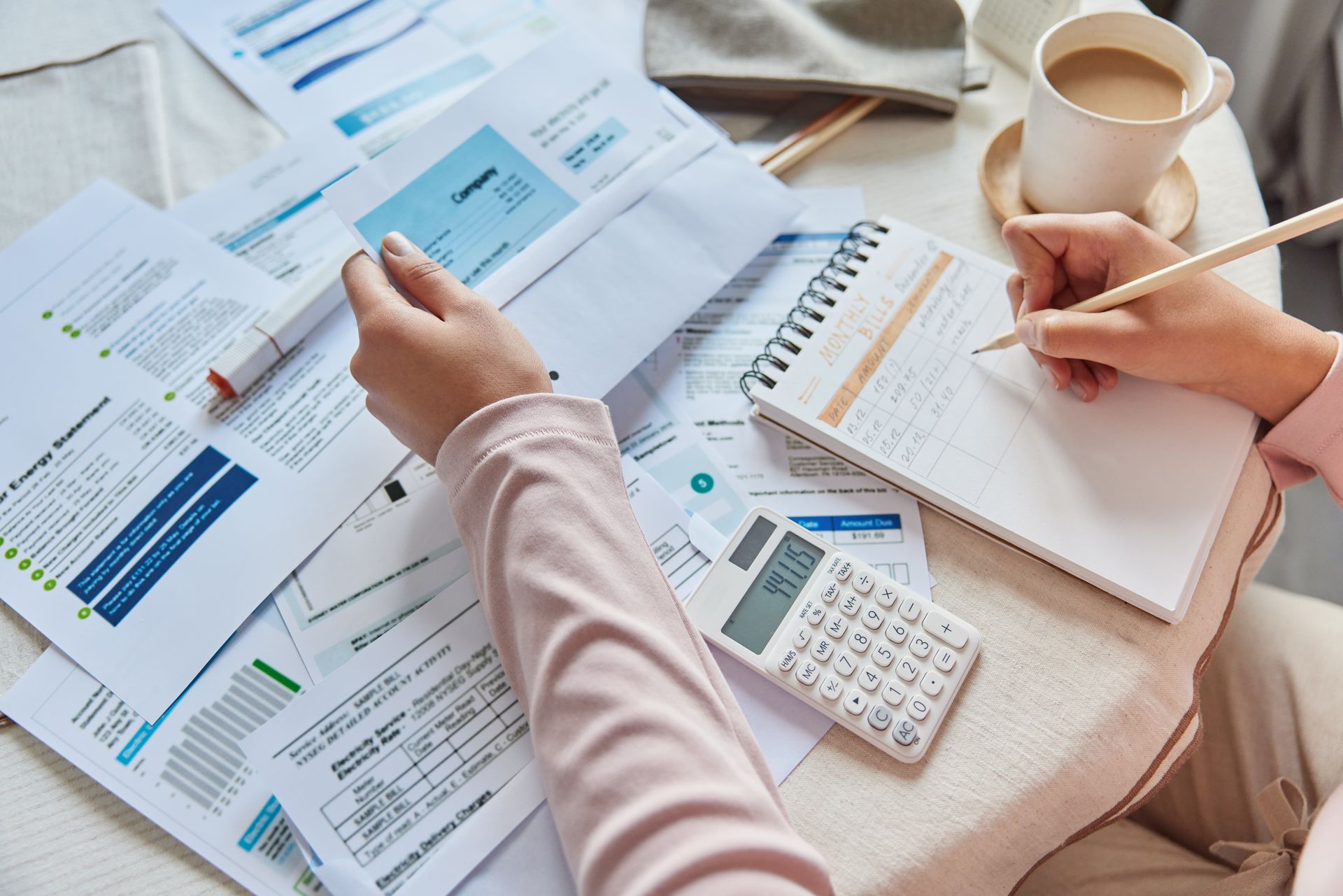 Person reviewing bills and writing notes, with calculator and coffee.