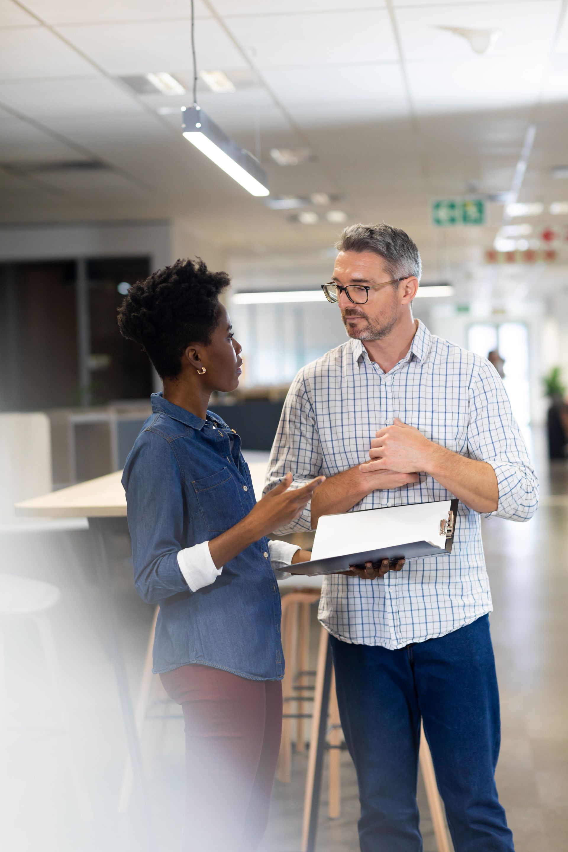 Woman and man in an office, discussing documents. Man holds tablet, both looking at each other.