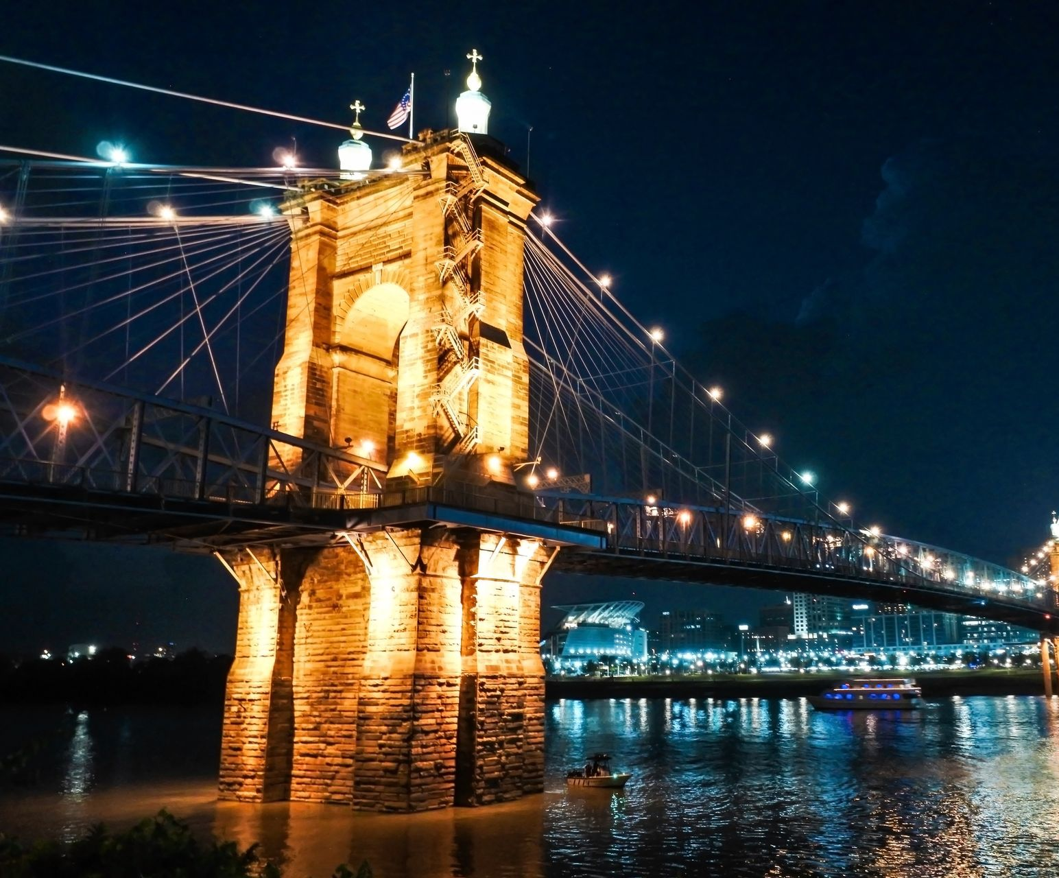 Suspension bridge lit up at night over a river, with cityscape visible in the background.