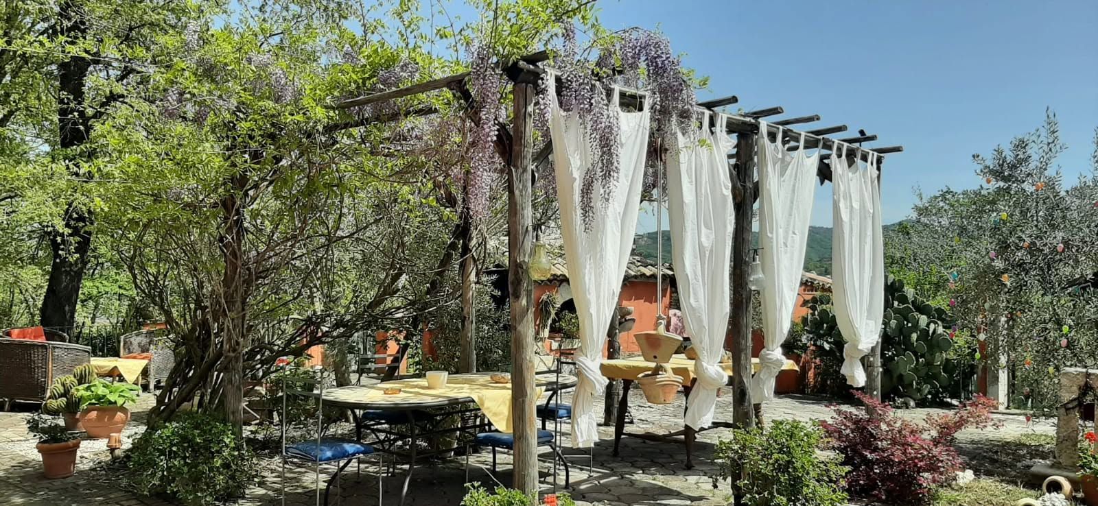Patio in giardino con tende di tessuto bianco che pendono da un pergolato tra alberi e piante in vaso.