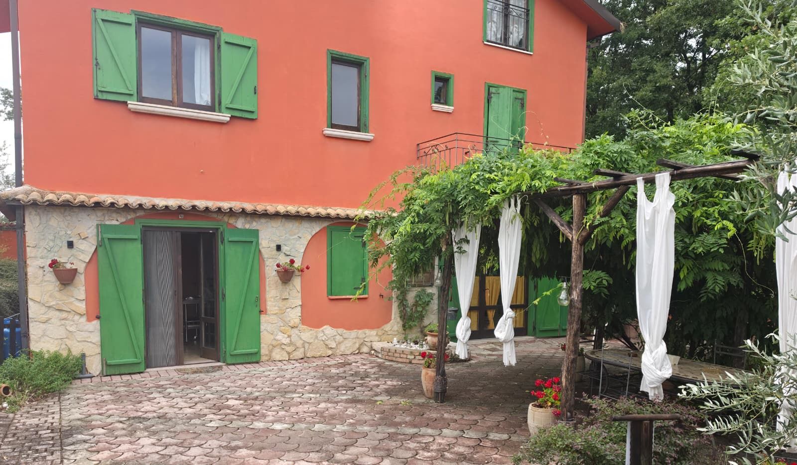 Casa rossa a due piani con persiane verdi e un pergolato ricoperto di viti sopra un patio in pietra.