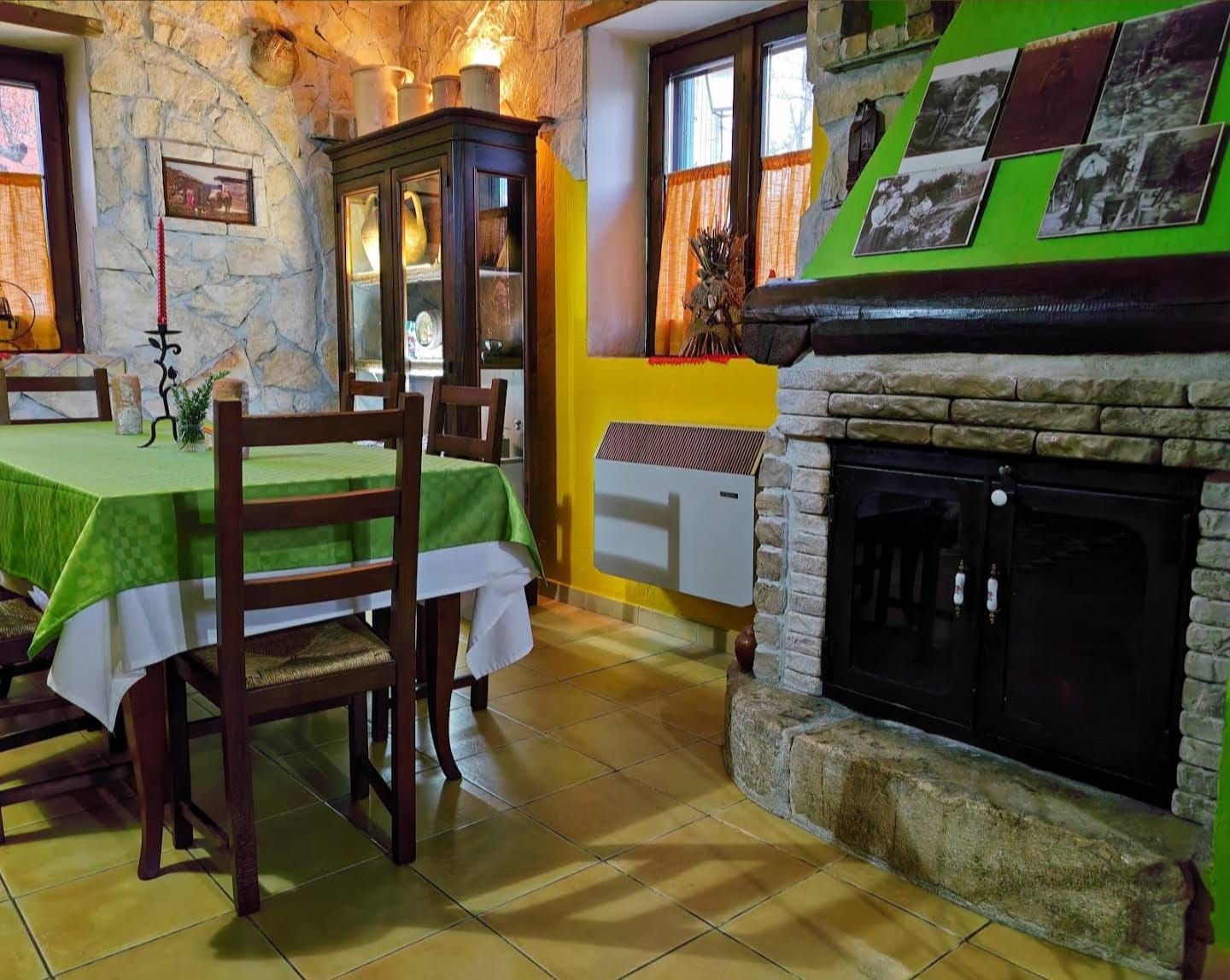 Accogliente sala da pranzo in stile rustico con tovaglia verde, sedie in legno, camino in pietra e luce naturale alla finestra.