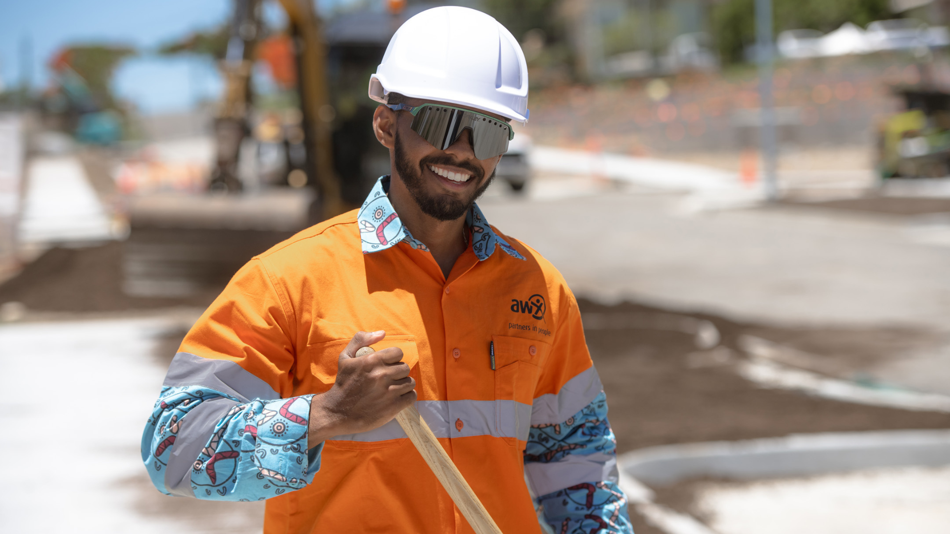 Three construction workers in orange safety gear on a sunny worksite. One holds a shovel, smiling.