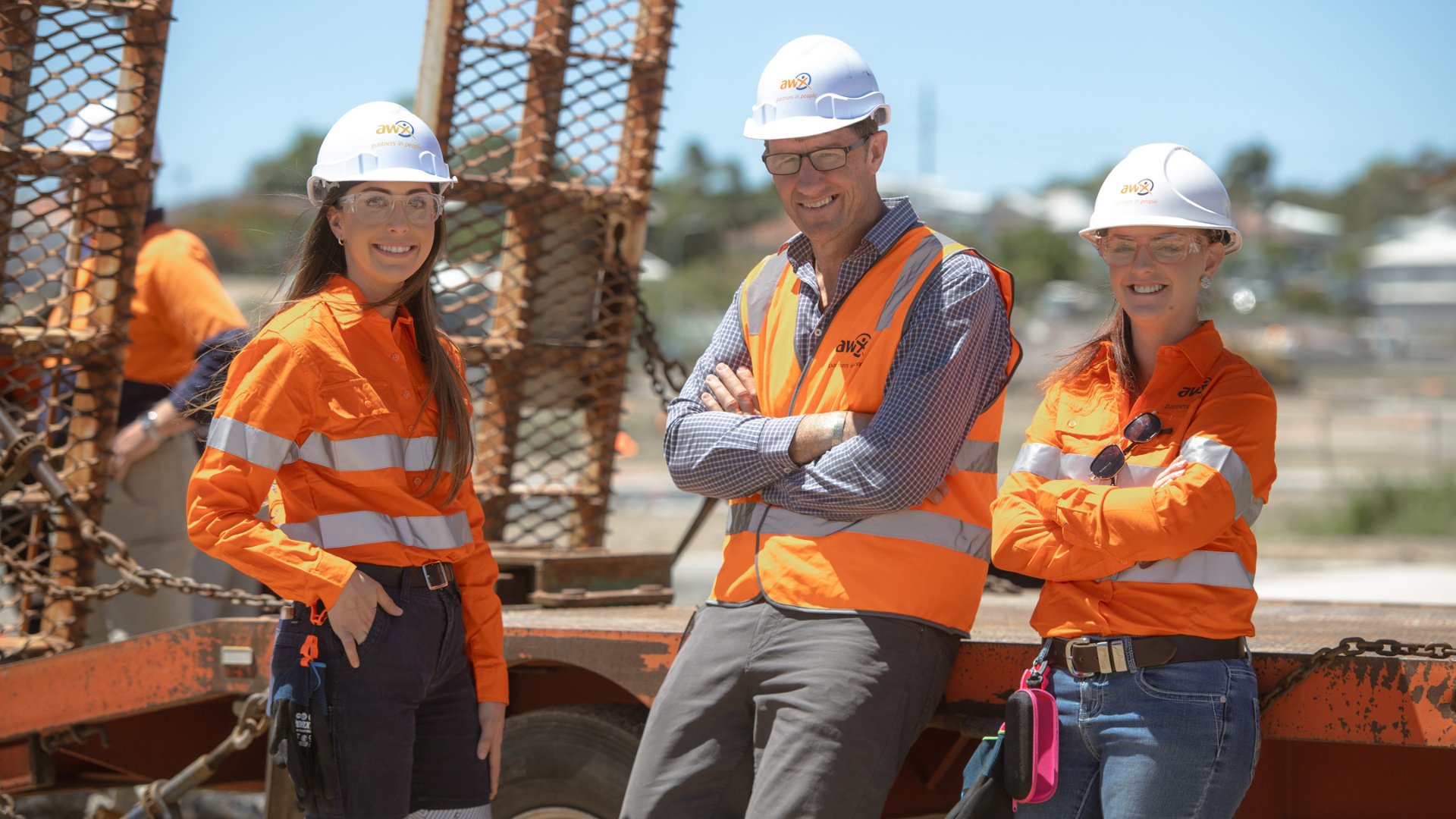 Three construction workers in orange safety gear and hard hats stand smiling at a worksite.