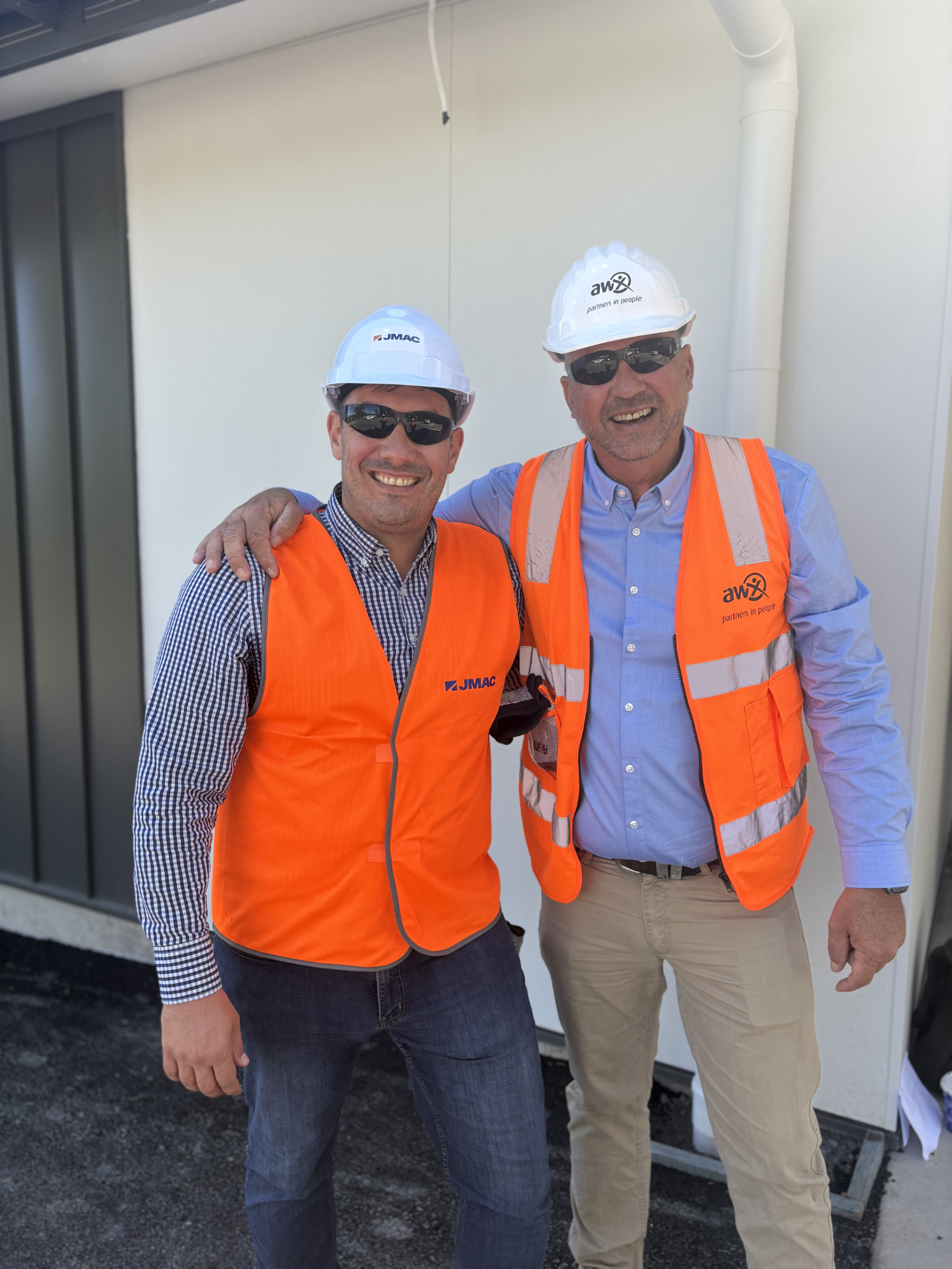 Two workers in hard hats and orange safety vests standing side by side outdoors, smiling.