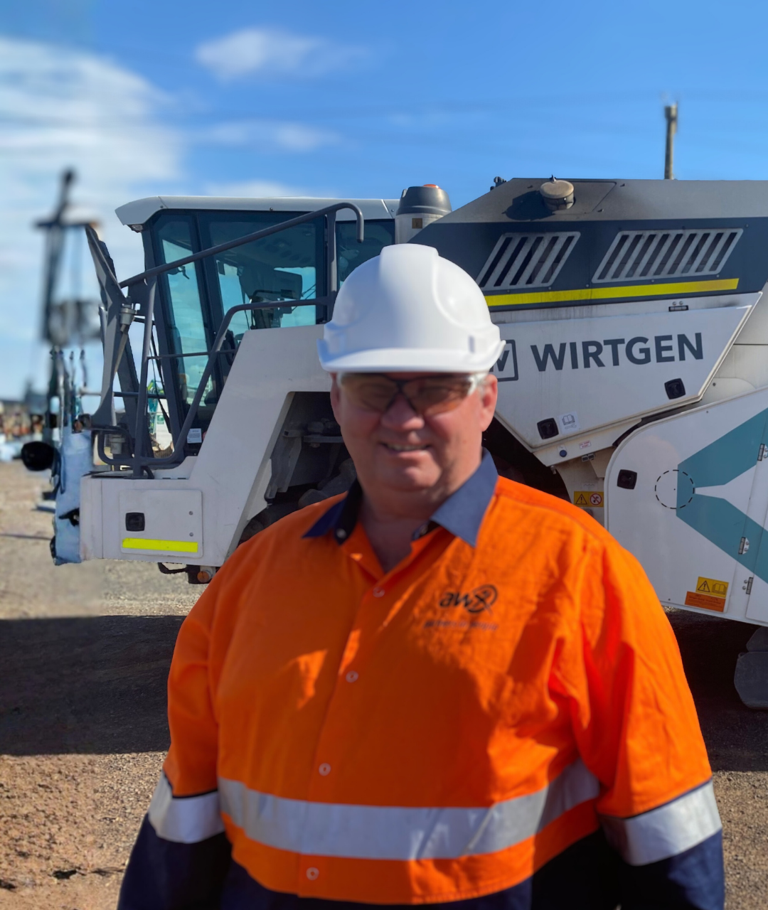 Man in orange workwear and hard hat, smiling, stands in front of a Wirtgen road milling machine.