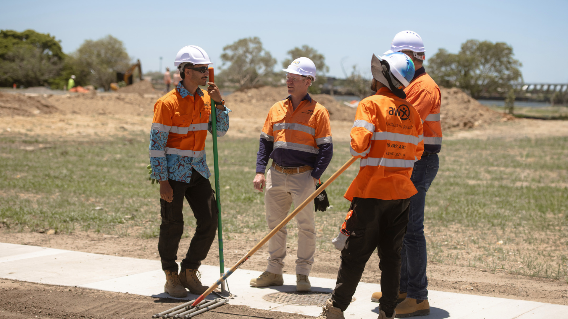 Four utility workers in orange safety gear and hard hats stand outdoors beside a pavement repair site.