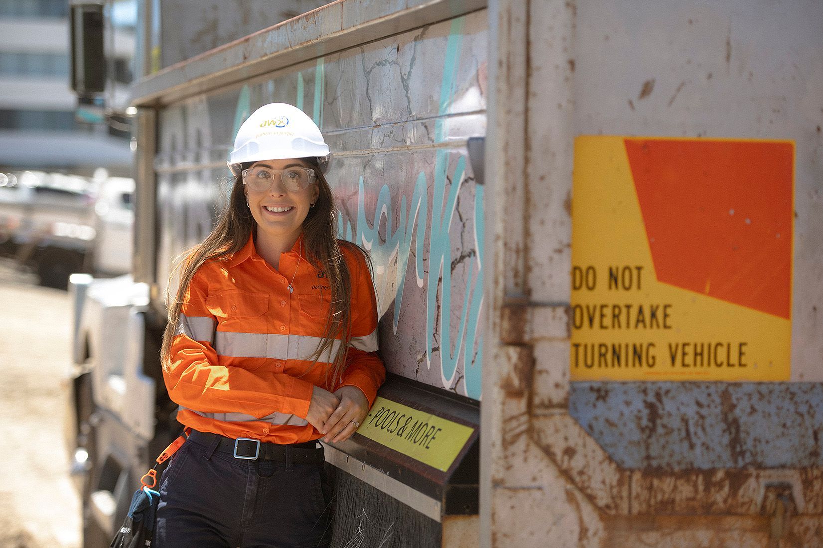 Worker in white hard hat and orange safety jacket standing by a truck with a “Do not overtake turning vehicle” sign
